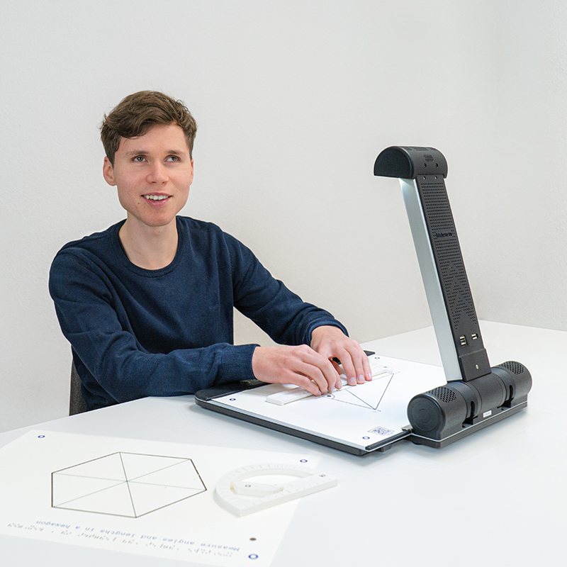 A young man sits in front of the Tactonom Reader and smiles. He is trying out the magic ruler together with the tactile graphic of a triangle on the Tactonom Reader Flex. Next to him is a smart protractor.