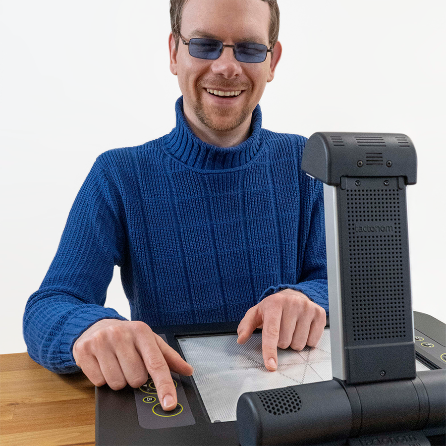 In front of the Tactonom Pro sits a smiling blind man, tapping the mathematical function displayed on the device with one finger and pressing the device’s confirmation button with the other finger.