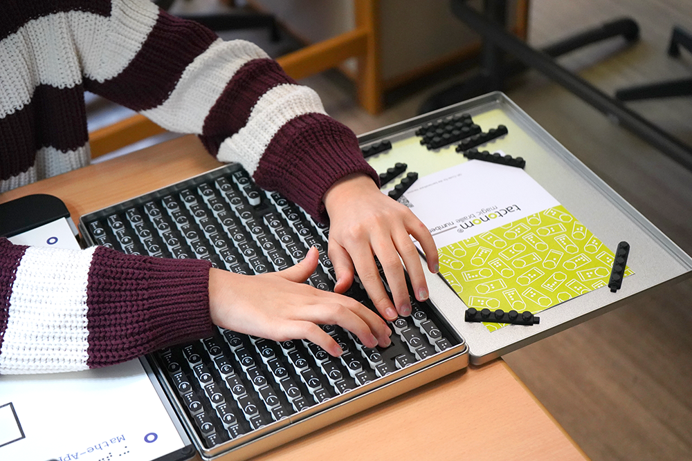 Die Box mit den Magic Braille Numbers, den magischen Zahlen, liegt offen auf dem Tisch. Man sieht die Hände einer Schülerin. Sie sucht nach bestimmten Zahlen und Rechen-Zeichen.