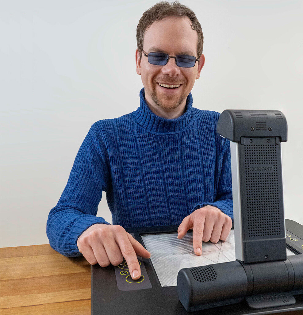 Unser blinder Mitarbeiter Martin sitzt vor dem Tactonom Pro und lächelt herzlich in Richtung Kamera. Der Zeigefinger seiner linken Hand liegt auf der Enter-Taste auf der rechten Seite des Gerätes. Der Zeigefinger der anderen Hand liegt auf der Grafikfläche auf. Die Pins bilden eine mathematische Funktion in einem Koordinatensystem.