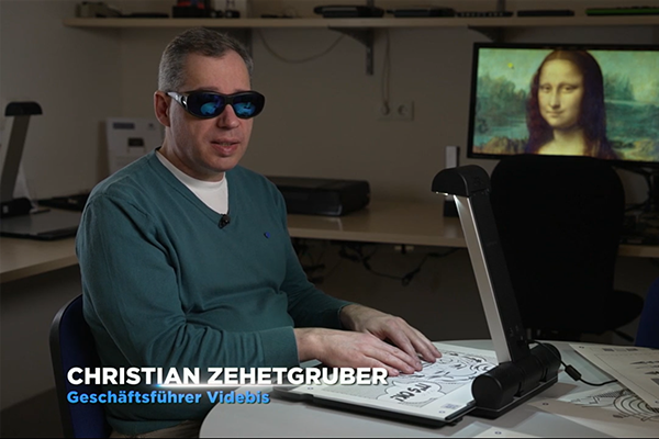 Christian Zehetgruber, Geschälftsführer von Videbis sitzt vor dem Tactonom Reader Flex auf dem eine Kunstgrafik im Pop Art Stil aufliegt. Es ist der Kopf einer Frau dargestellt.