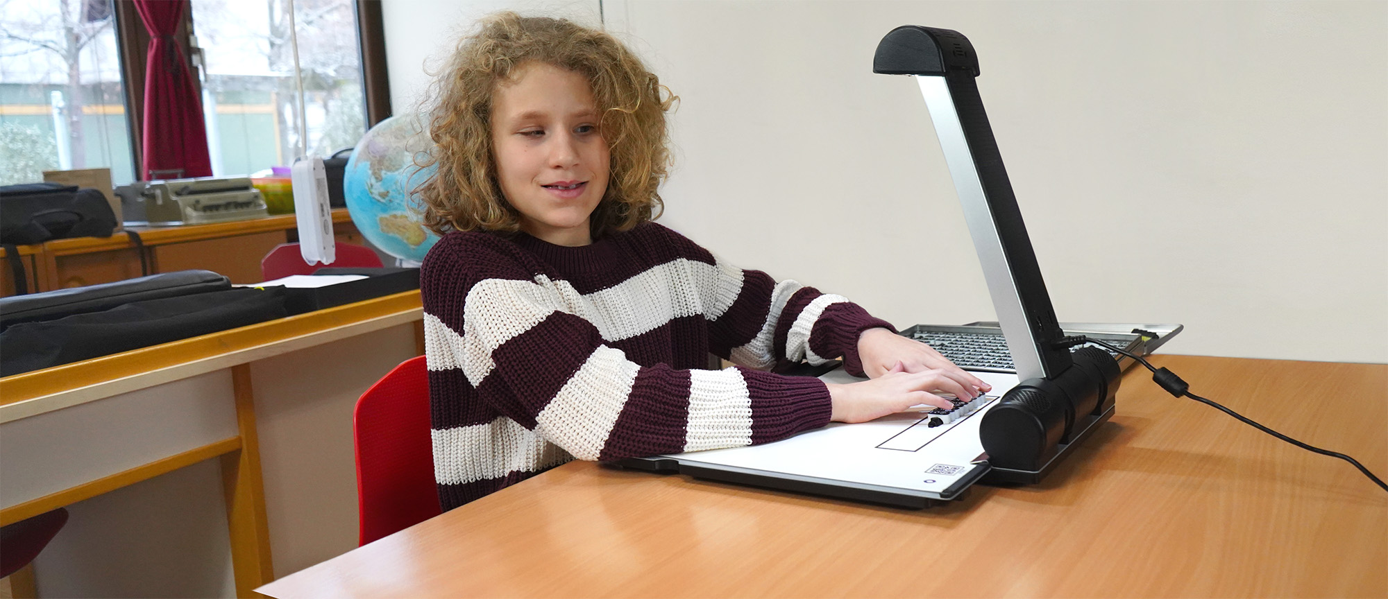 Eine Schülerin mit blonden Locken und rot-weißem Streifen-Pulli sitzt vor dem Tactonom Reader Flex im Klassenzimmer.